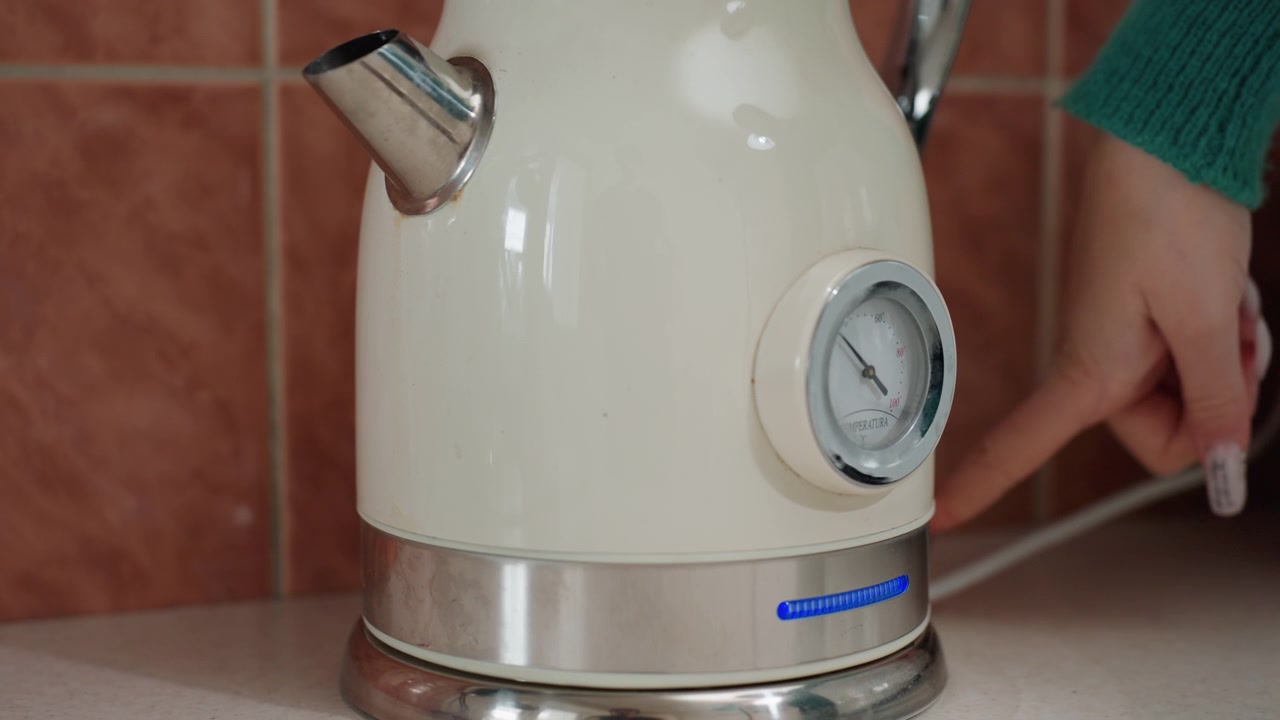 Bollitore color crema sul bancone della cucina con piastrelle, primo piano mostra base, coperchio, posizionamento, mano che si avvicina all'interruttore, indicatore blu in attesa di attivazione, accenti cromati retrò e manometro suggeriscono un elettrodomestico vintage.