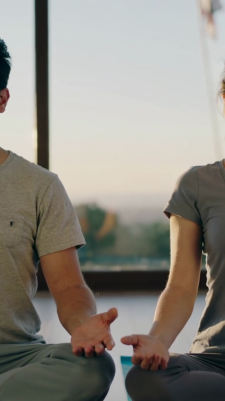 Vertical video: Sitting couple meditating at home on yoga mats by large window, wearing grey tees