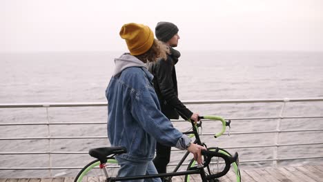 Schönes-Lächelndes-Paar-Junger-Hipster,-Die-Am-Herbsttag-Zusammen-Mit-Ihren-Fahrrädern-Am-Meer-Spazieren-Gehen