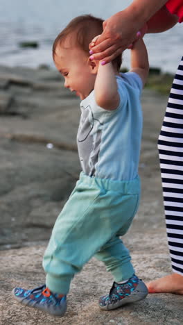 cute little baby studying to walk at the rocky shore. mommy is holding her son's hands. water at backdrop. vertical video