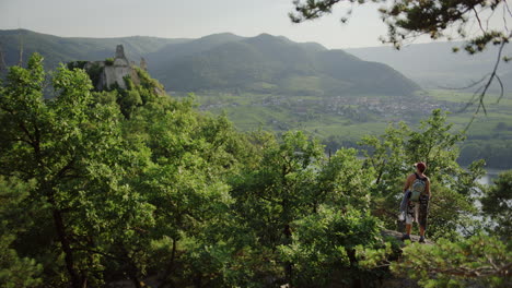 hiker-looking-over-the-valley,-seeing-durnstein-castle,-burgruine,-green-mountain-Austria-horizon,-athletic-outdoor-hiking-man-on-a-trekking-trail-walk-in-nature