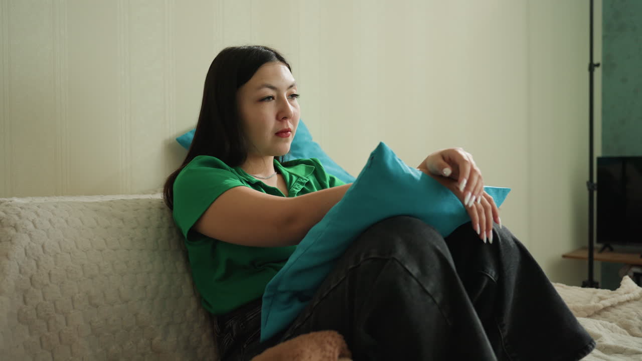 소파에 앉아 쿠션을 끌어안고 있는 여성이 생각에 잠긴 자세로 먼 곳을 응시하고 있어요. 차분한 실내 톤, 창의적인 성찰과 조용한 휴식을 암시하는 고요한 일상의 순간입니다.