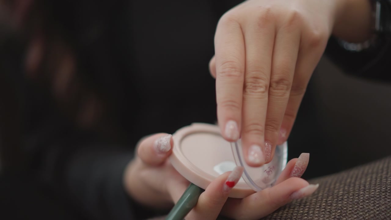 Tight close view of hands opening round cosmetic compact while steadying case with other hand and holding small rolled paper or thin stick, showing careful lid movement in warm indoor light