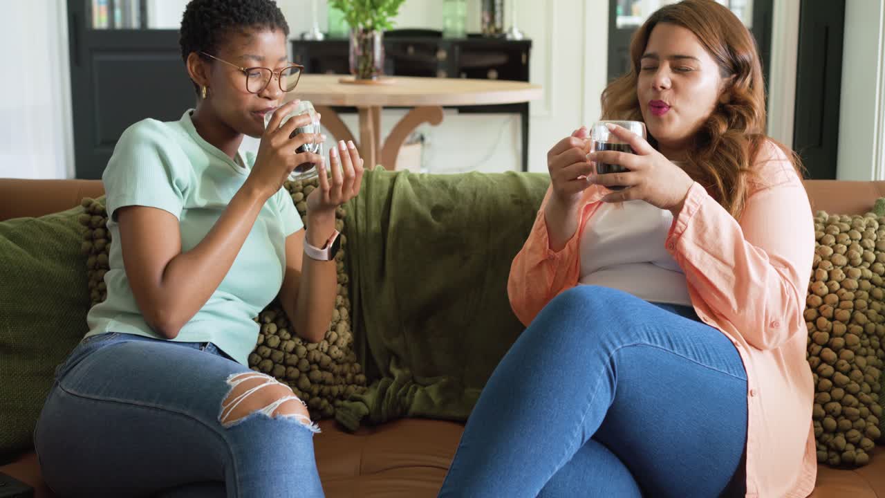Diverse female friends holding glass mugs on couch smelling and sipping in living room, tasting
