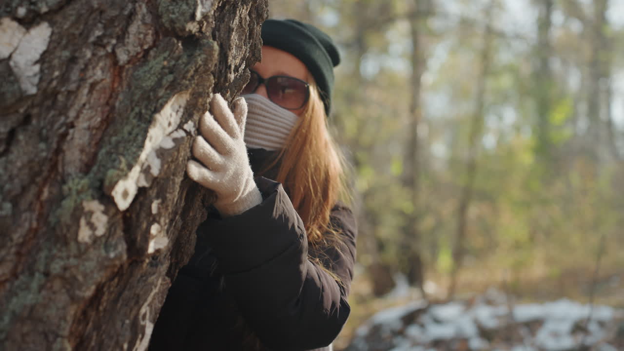 White woman masked behind tree scouting, snowy woodland scene with sunglasses and knit beanie, gloved hand gripping bark, furtive prowler movement, tense suspenseful mood, watchful gaze peeking