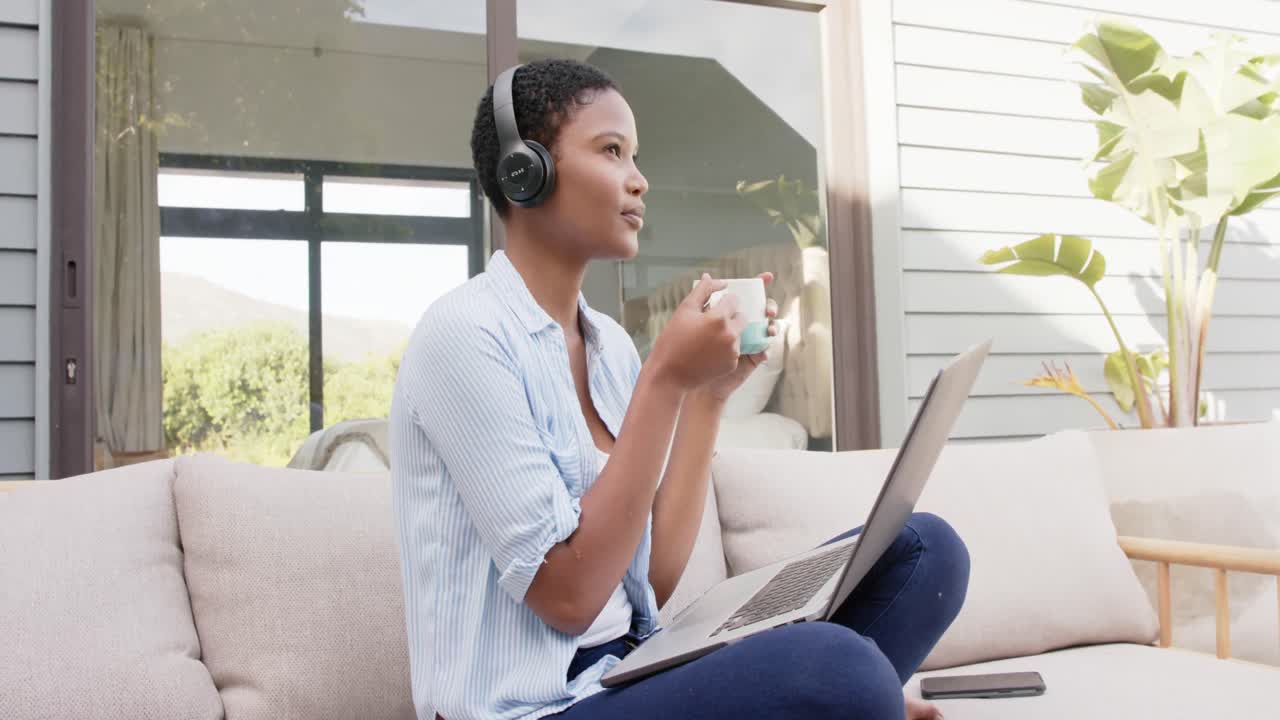 African american woman sitting on sofa on terrace wearing headphones, using laptop, slow motion