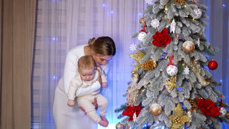 woman wearing white dress holds a baby standing near the christmas tree. mother kick one of the balls on the tree with kid's foot.