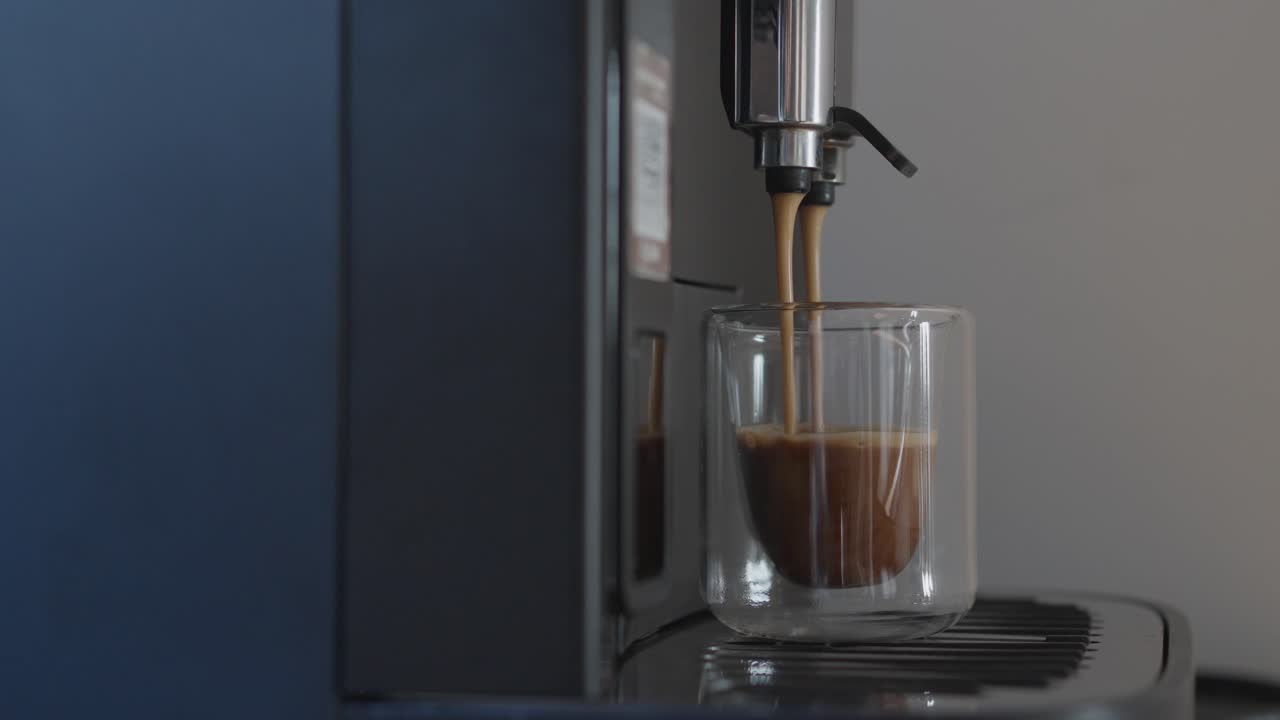 Nærbilde av fersk espresso som helles fra en kaffemaskin ned i en gjennomsiktig glasskopp, med fokus på crema, tekstur og myke bevegelser i et rent innemiljø