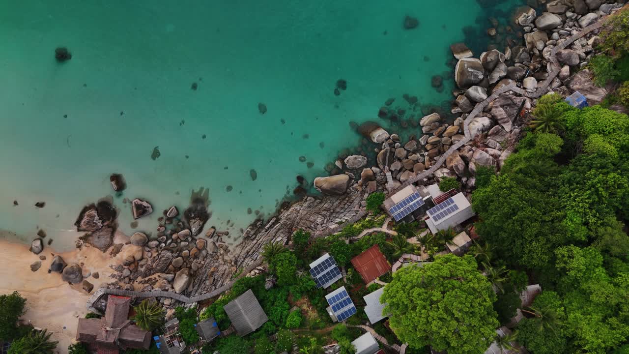 Top-down aerial footage of eco resort with solar panels, rocky coastline, sandy beach, and turquoise sea in Thailand. Perfect for eco-tourism, sustainable travel, nature, and adventure projects.
