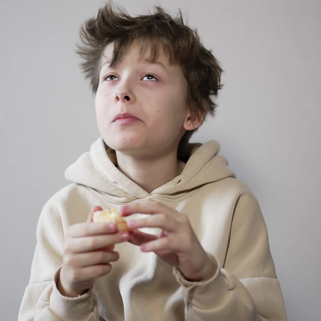 Calm brunet teenager eating a tangerine. Boy with messy hair eats fruit looking around and then into camera