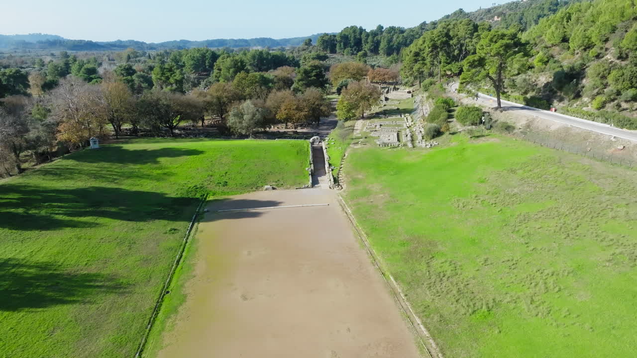 Ground-level path runs alongside ancient Olympia ruins in green, tranquil area, aerial establishing