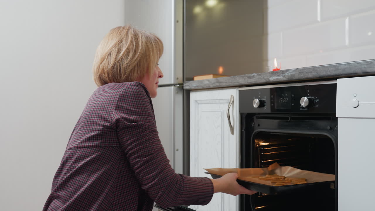 Donna di mezza età che apre lo sportello del forno, inserendo dei biscotti su una teglia, frigorifero che riflette la luce in una cucina moderna, preparando prodotti da forno in un ambiente accogliente e ben illuminato