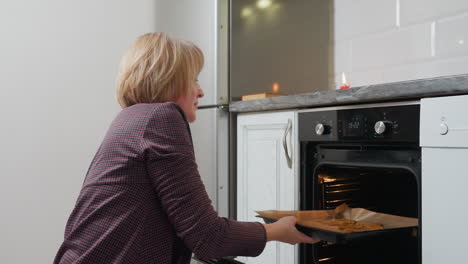 middle-aged woman opening oven door, placing cookies inside on tray, standing fridge reflecting light in modern kitchen, preparing baked goods in cozy, well-lit kitchen environment