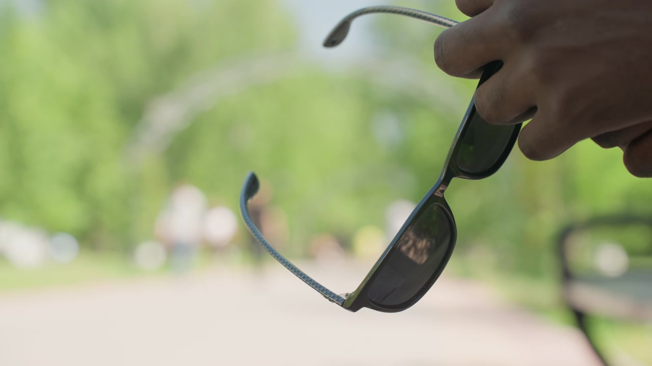 Close up of hand holding sunglasses with dark lenses and blue arms against outdoor park background. scene shows soft natural light reflecting on lens emphasizing texture detail in both
