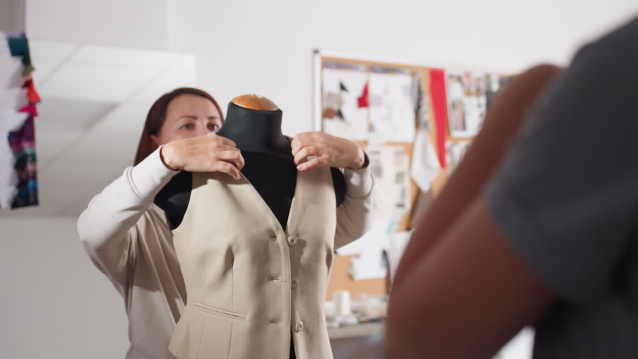 Fashion designer slips beige sleeveless blazer off black dress form, checking neckline and shoulder fit while smiling at colleague in bright studio, celebrating precise tailoring milestone