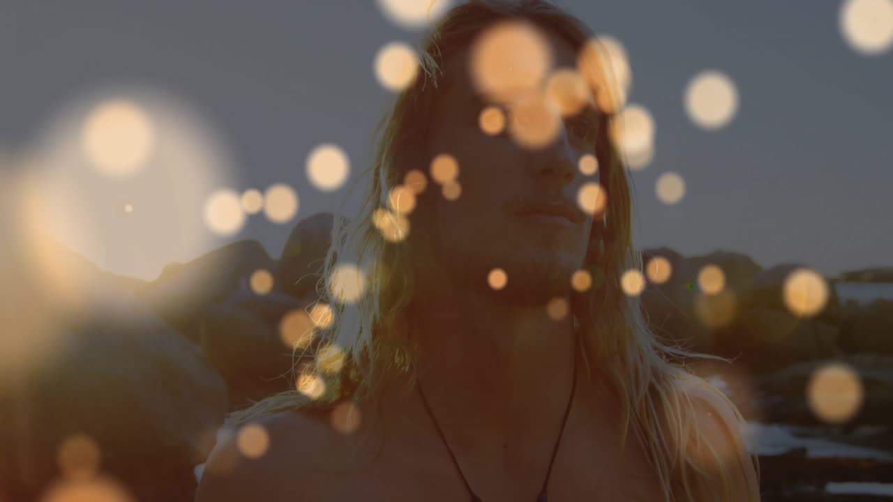 Videografo che riprende una modella che gira la testa sulla costa al tramonto, con luci bokeh fluttuanti che rivelano la fotocamera.