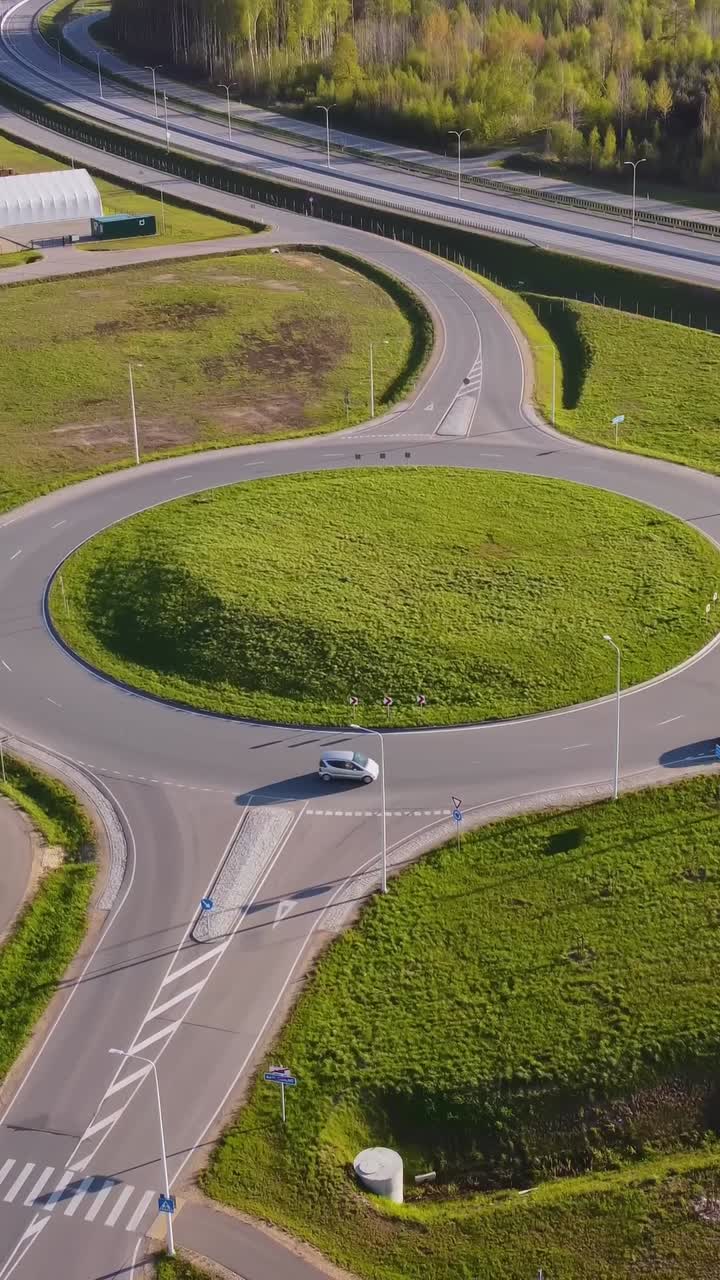 Vertical drone static view shows Katlakalns roundabout with grassy central mound, cars circulating, adjacent access road, distant highway, and pine forest bordering the scene under afternoon light