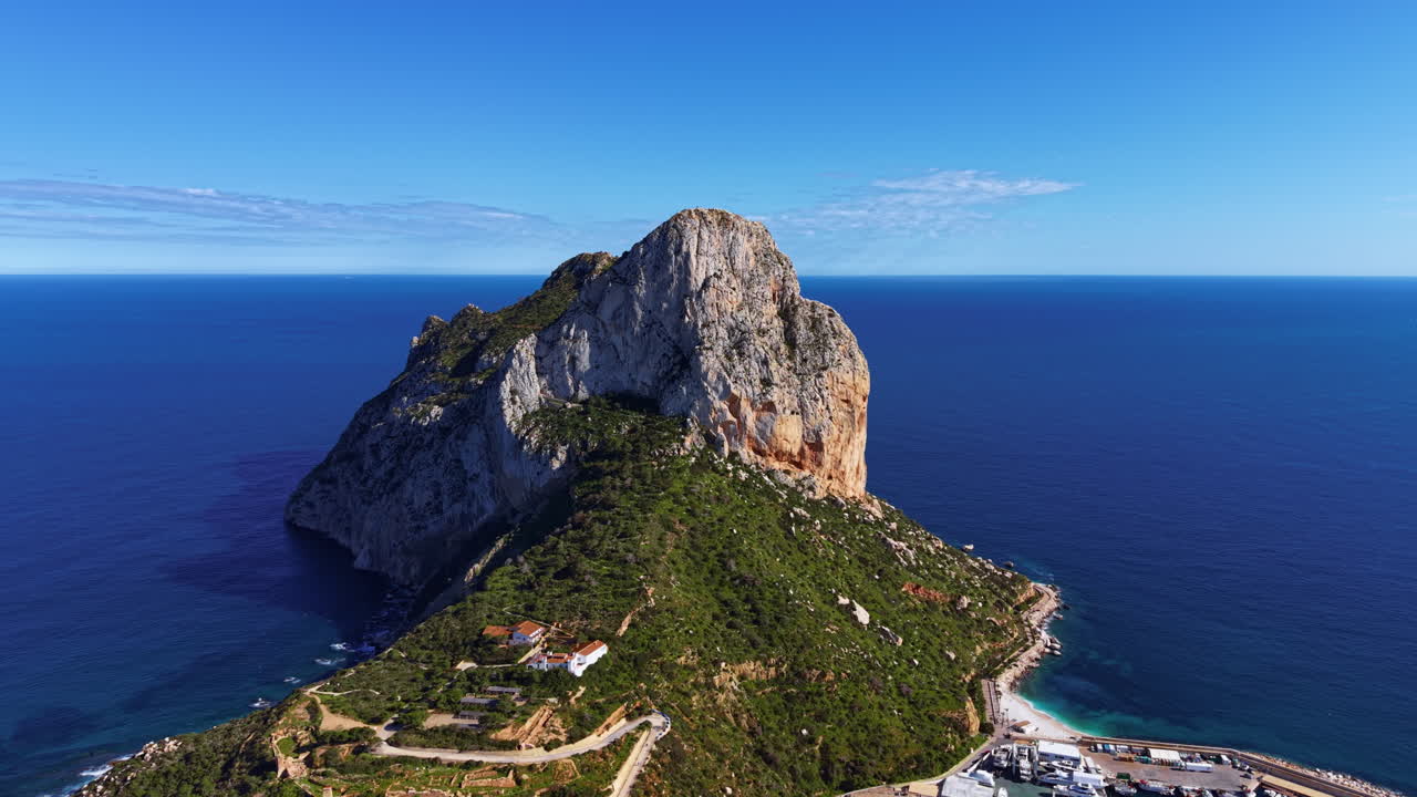 Formazione rocciosa Penon de Ifach che si erge drammaticamente sul Mar Mediterraneo vicino a Calpe, Spagna, con un piccolo porto ed edifici costieri visibili alla sua base.