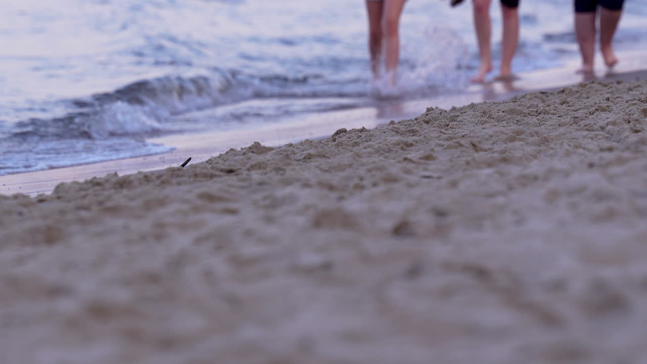 Group of people walking barefoot by the sea, waves touching sandy beach at sunset