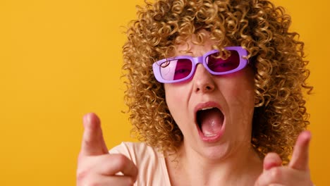 happy optimistic young woman in stylish veri pery color glasses raises arms and dances to favorite music, forgot about all problems isolated over yellow background