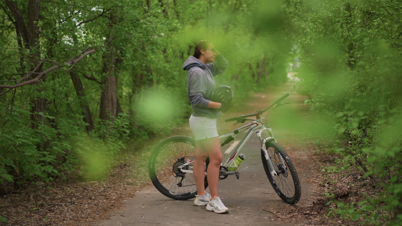 Asian Cyclist Pausing With Bike On TreeLined Path, Helmet On And Hand Near Ear As If Checking Route On Phone, Contemplative Stance Under Green Tunnel, Preparing To Continue Journey