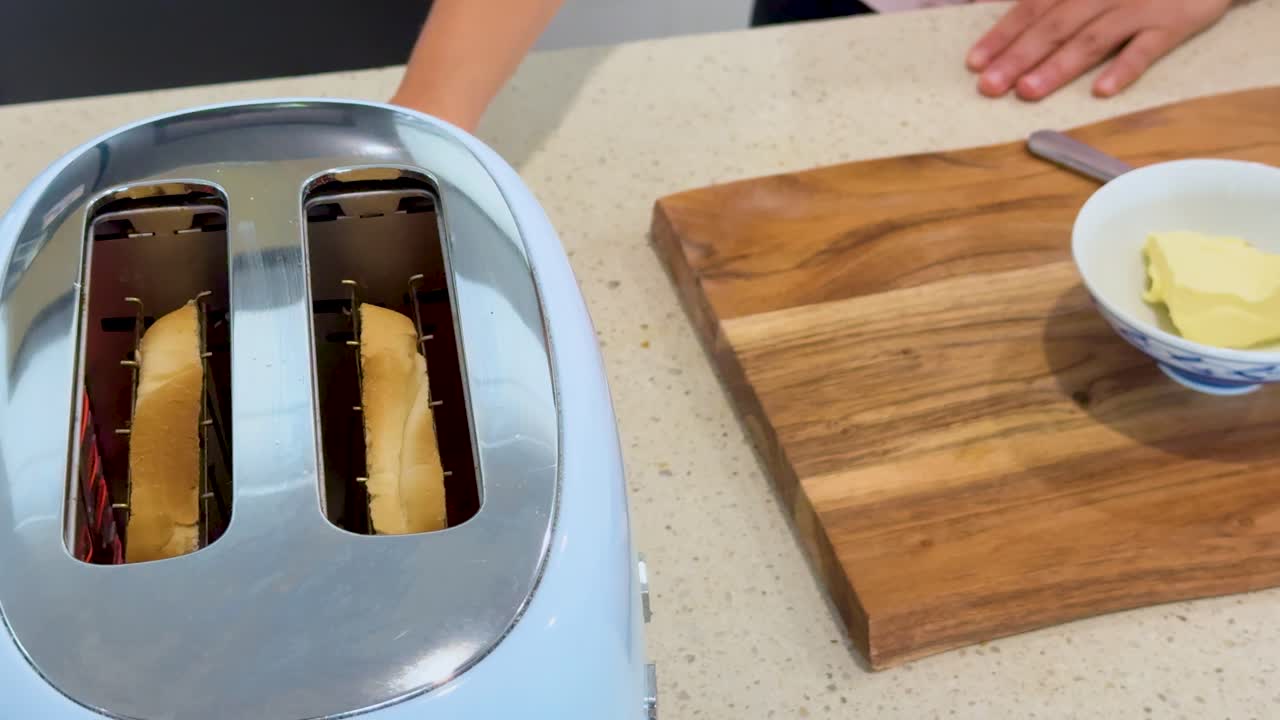 Person waits beside toaster as bread slices brown, readying butter and knife on wooden board under bright, even kitchen lighting. Static camera, close-up view