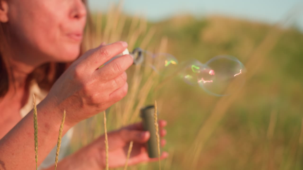 Close up young woman gently blowing iridescent soap bubbles over sunlit grassy hill at sunset, vibrant rainbow reflections drifting into warm breeze against blurred nature