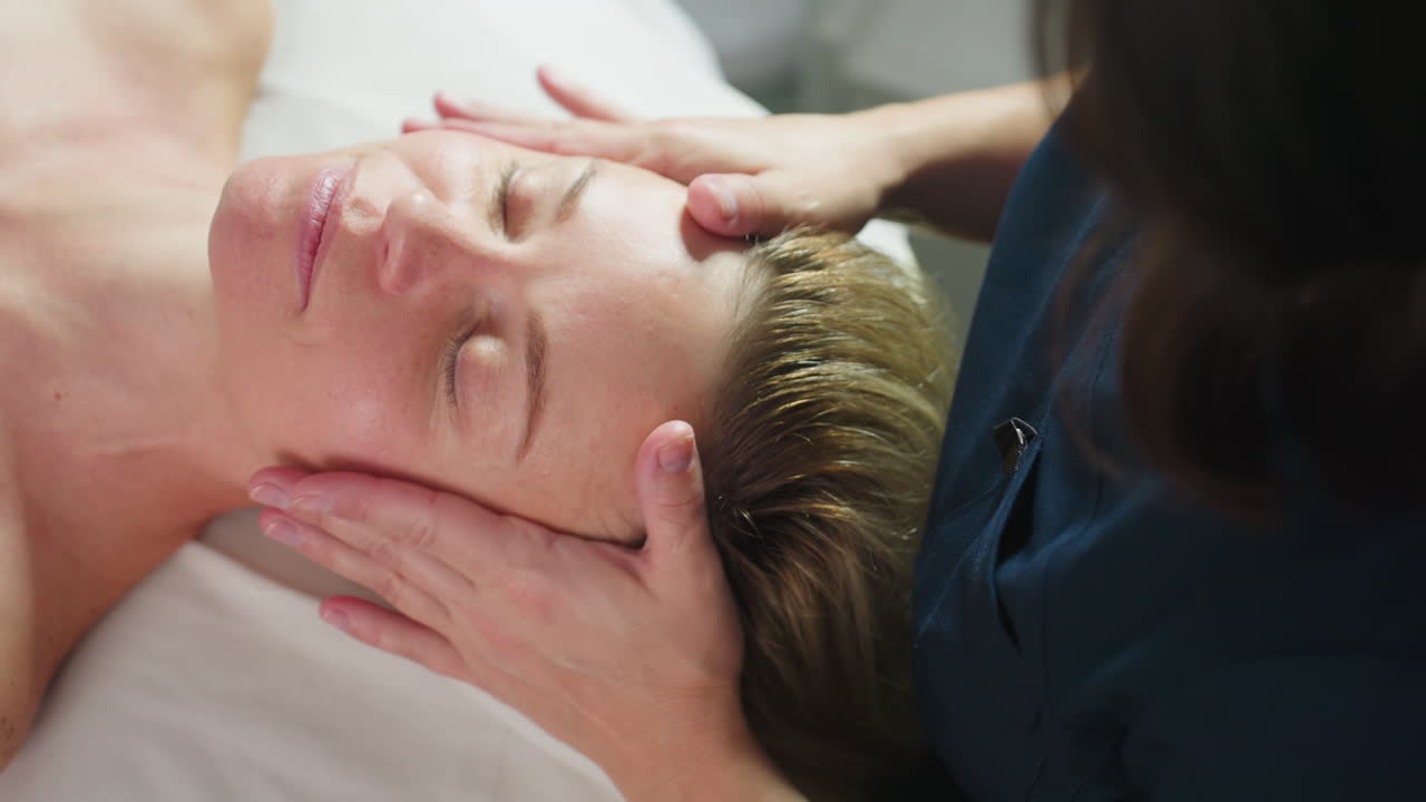 Wellness technician massaging female client head and neck while client closes her eyes softly on massage chair in spa with white background during tranquil therapy session showcasing expert touch
