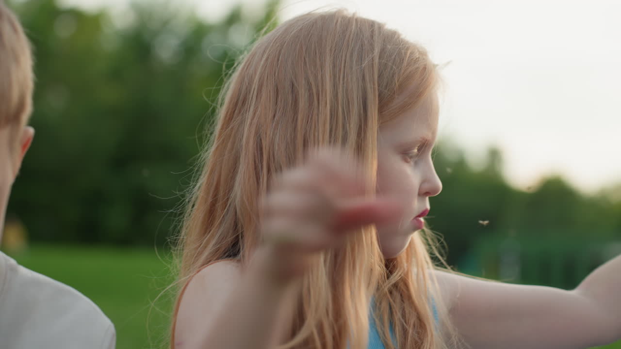 young girl chatting with sibling while putting on picnic jacket, seated on grass during relaxed park outing, soft evening light, casual play, close interaction showing care and companionship