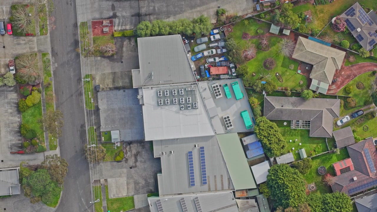 Dynamisk dronevideo af et datacenter med solpaneler på taget, køleenheder og parkering, omgivet af grønne beboelsesområder i klart dagslys. Levende vidvinkeludsigt.