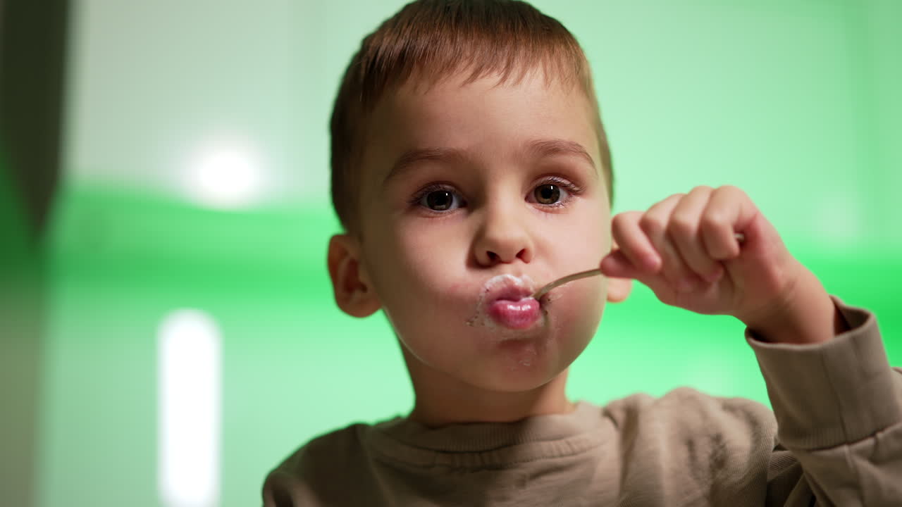 Face of a lovely Caucasian toddler holding a spoon in his mouth. Close up. Snacking time. Blurred backdrop.