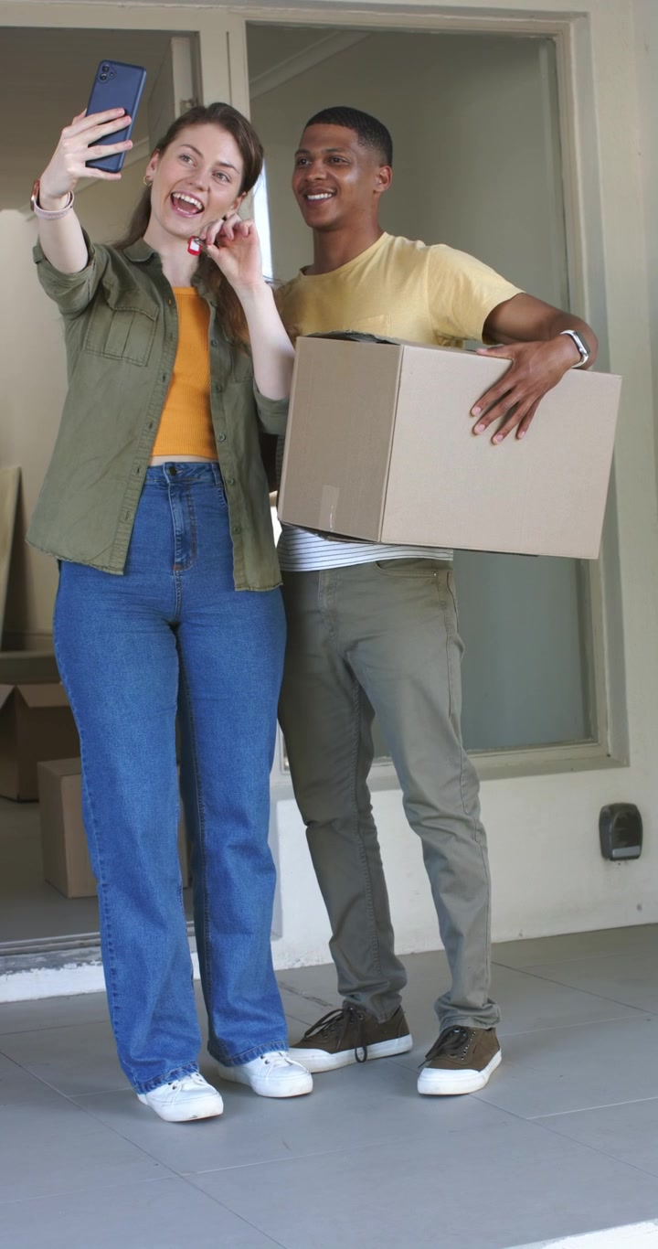 Vertical video: Diverse couple celebrating moving day by filming keys with smartphone at entrance