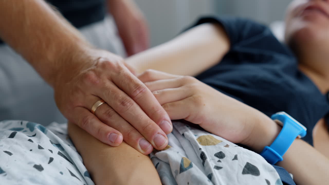 Male hand with wedding ring holding female one. Husband supporting pregnant wife lying in hospital bed. Blurred backdrop.