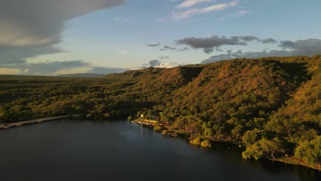 Schöne-Drohnenaufnahme-Von-Grünen-Wachsenden-Hügeln-Neben-Dem-Wassersee-Am-Abend