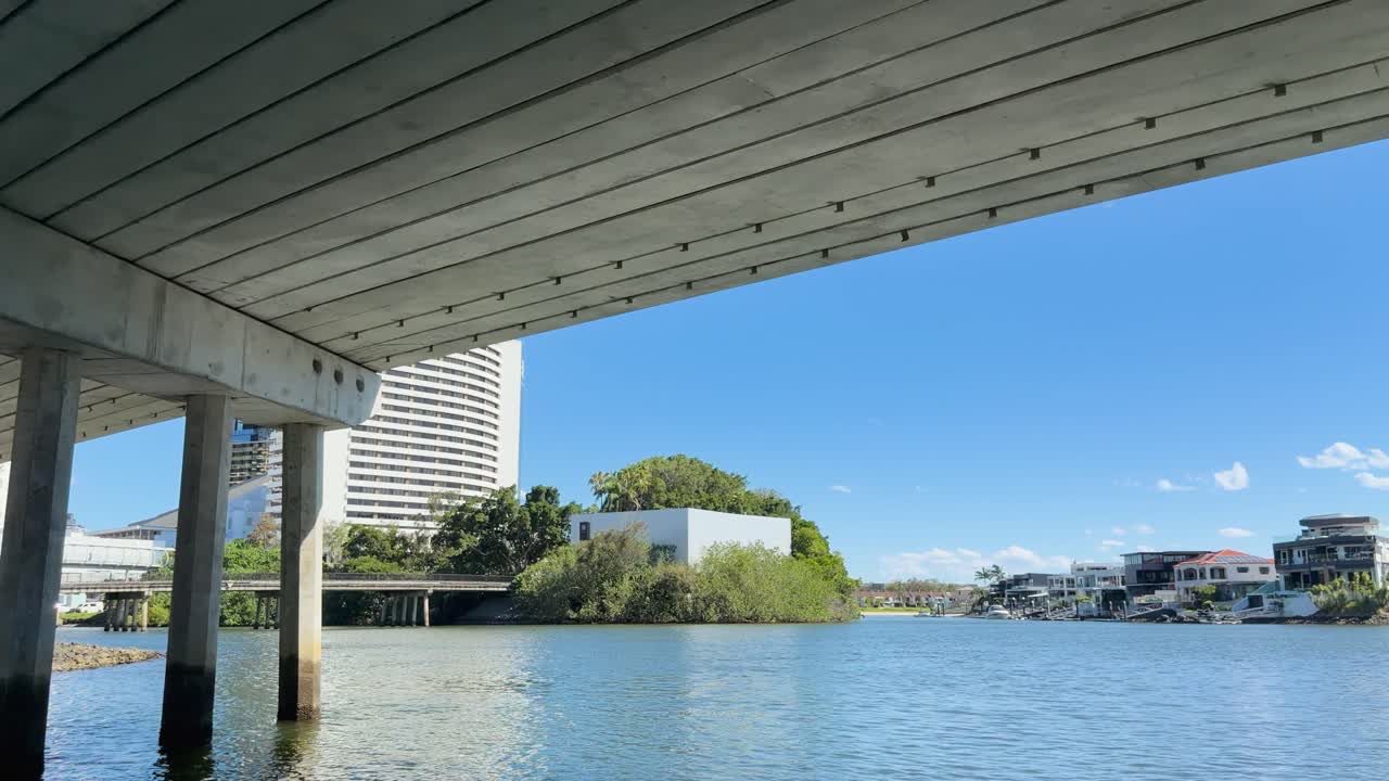 Camera glides beneath concrete bridge over river, revealing city buildings, blue sky, and water reflections