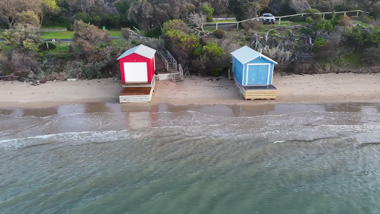 Drone glides above vibrant beach huts, sandy shoreline, and calm water in soft daylight