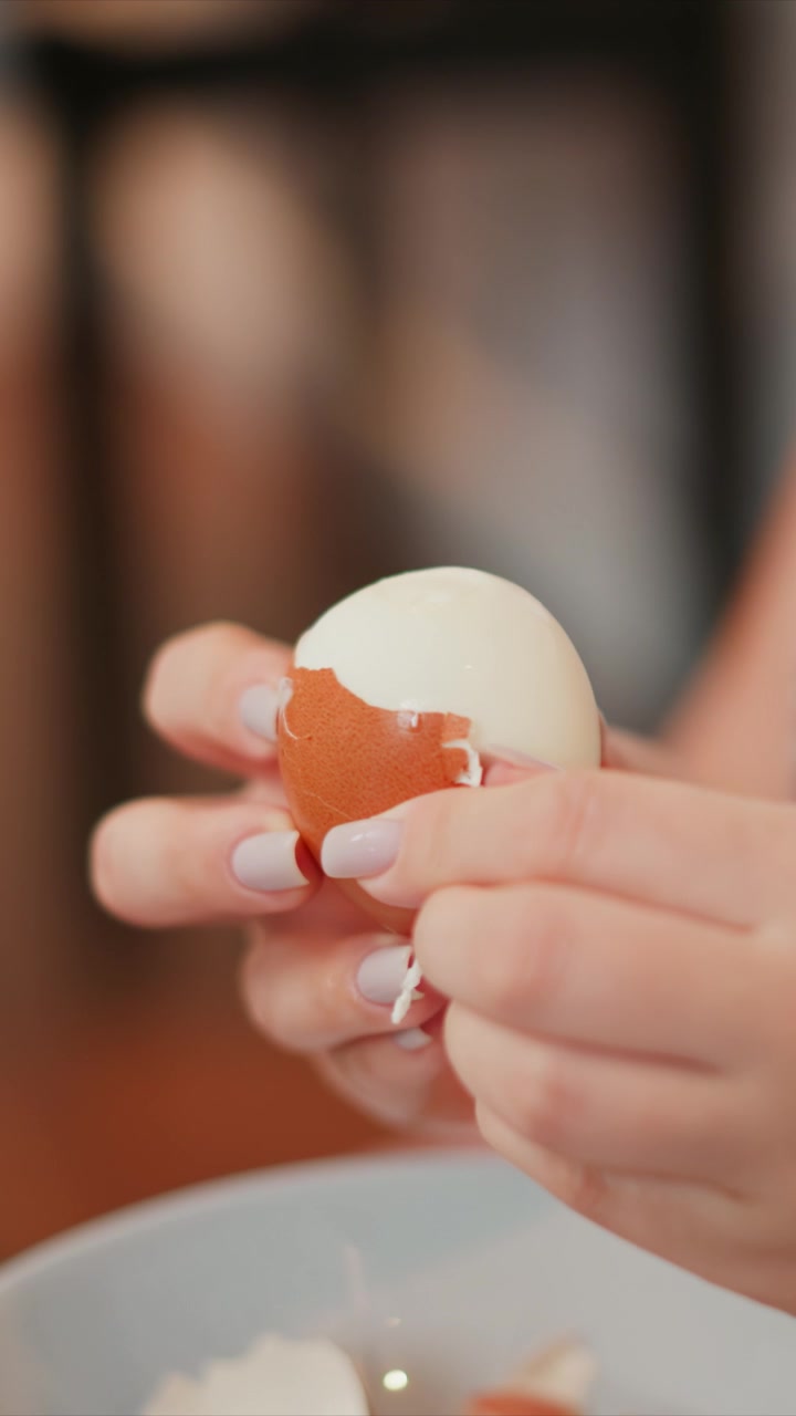 Woman carefully peels brown egg revealing white interior. hands with neatly manicured nails work steadily to remove shell fragments over soft lighting and out of focus background. bowl