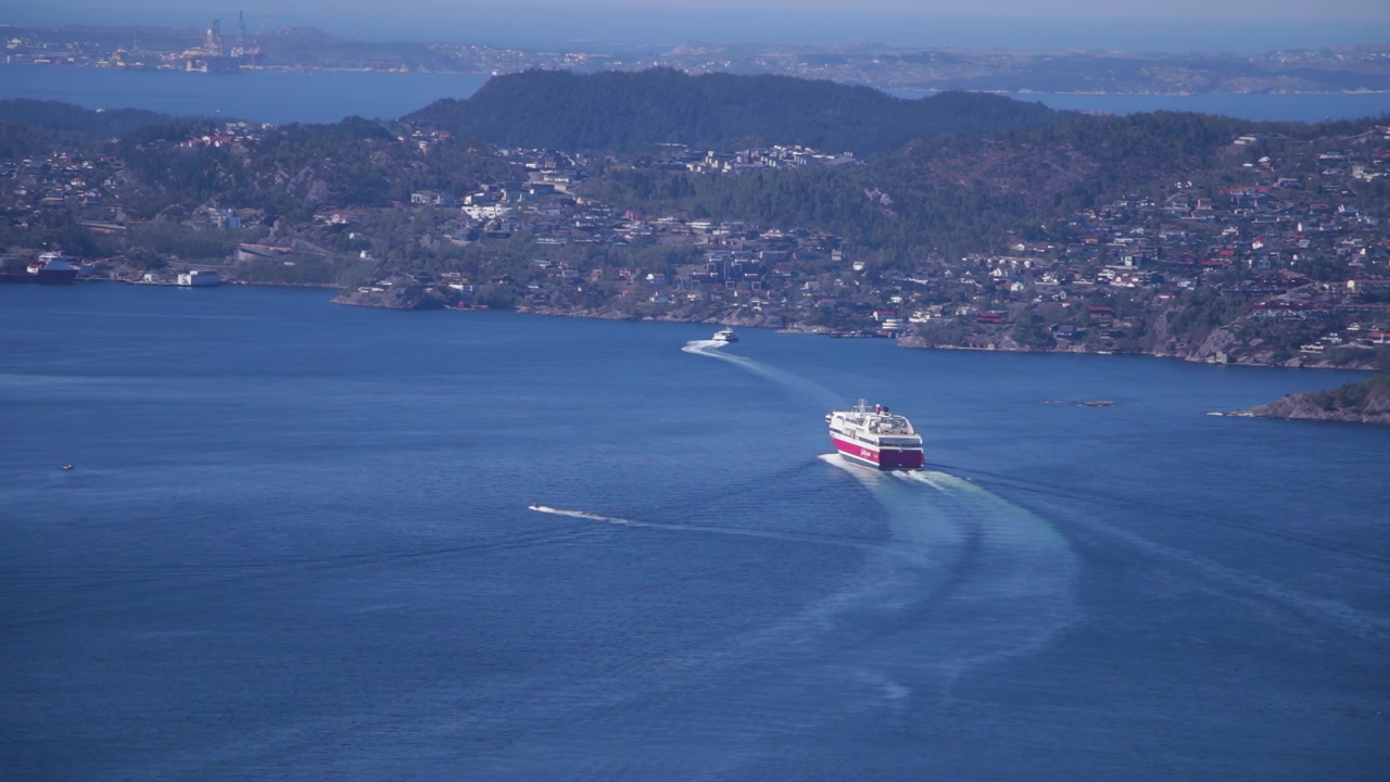 Stor færge og både sejler over blå fjord med Bergens forstæder i baggrunden
