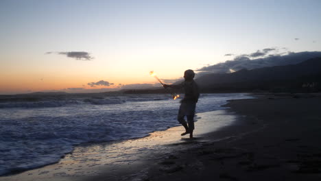 un hombre en silueta bailando y girando un bastón de fuego en llamas en la playa al atardecer con llamas y olas del océano en la orilla en cámara lenta