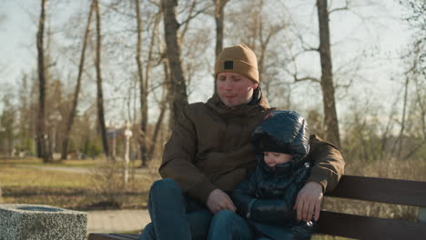 close up of father and son sitting close on a bench together smiling with the son folding his hand and the fathers leg crossed