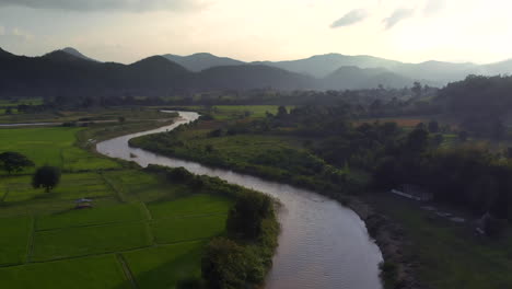 Vuelo-Aéreo-Sobre-El-Río-Y-Los-Campos-De-Arroz-Contra-Las-Montañas-En-El-Horizonte-Durante-La-Puesta-De-Sol