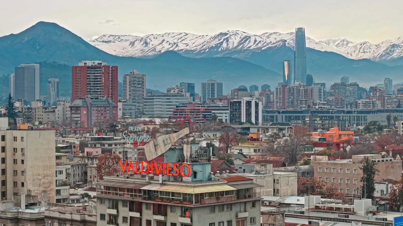Orbita aerea che rivela lo skyline di Santiago, Cile, con torri ed edifici moderni e le Ande sullo sfondo; insegna Valdivieso