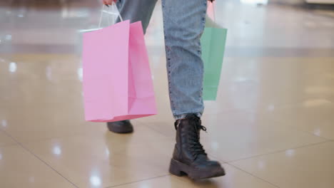 leg view of person wearing black boots and jeans, walking confidently through a modern mall with shopping bags in hand, sleek tiles on floor with blurred background