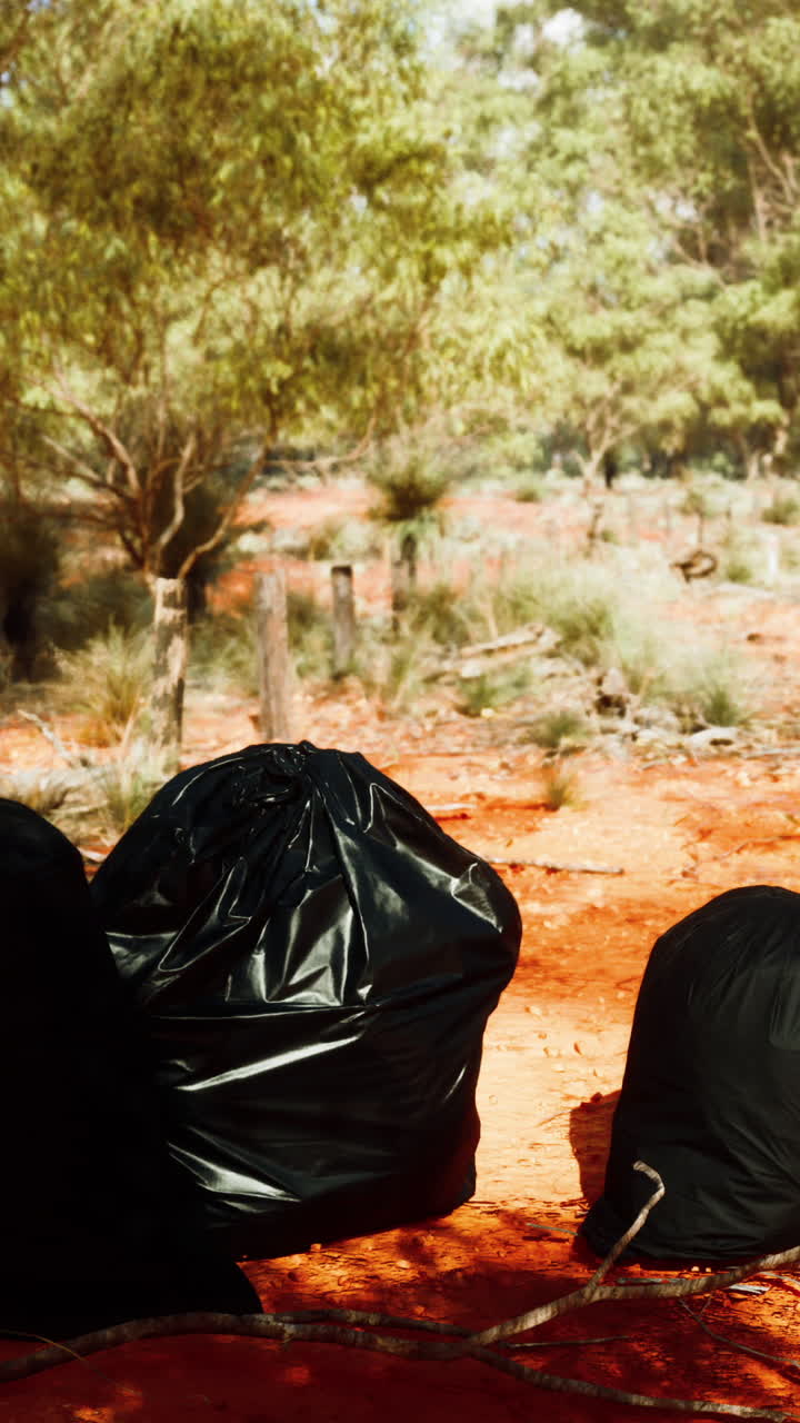 Three black trash bags sit on a red dirt path in the bush.