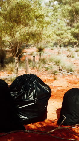 three black trash bags sit on a red dirt path in the bush.