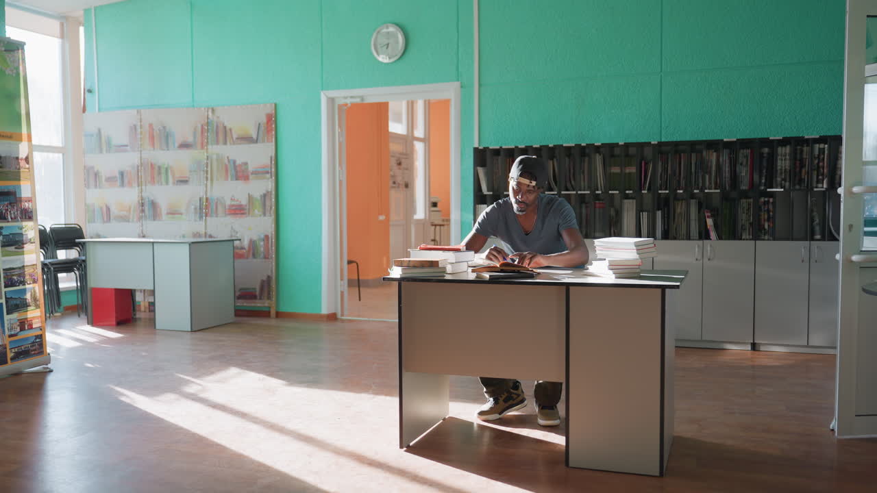 College boy sits at desk in bright library, surrounded by stacks of books, writing intently in notebook with pen, deeply focused on studies as sunlight streams across polished floor