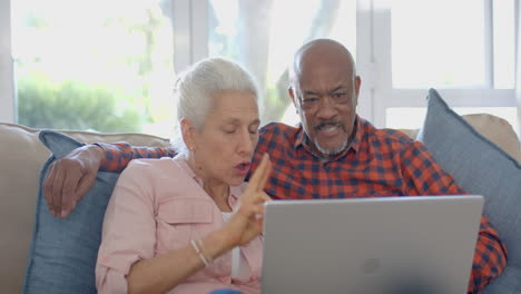 Happy-senior-biracial-couple-sitting-on-couch-and-using-laptop-at-home,-slow-motion