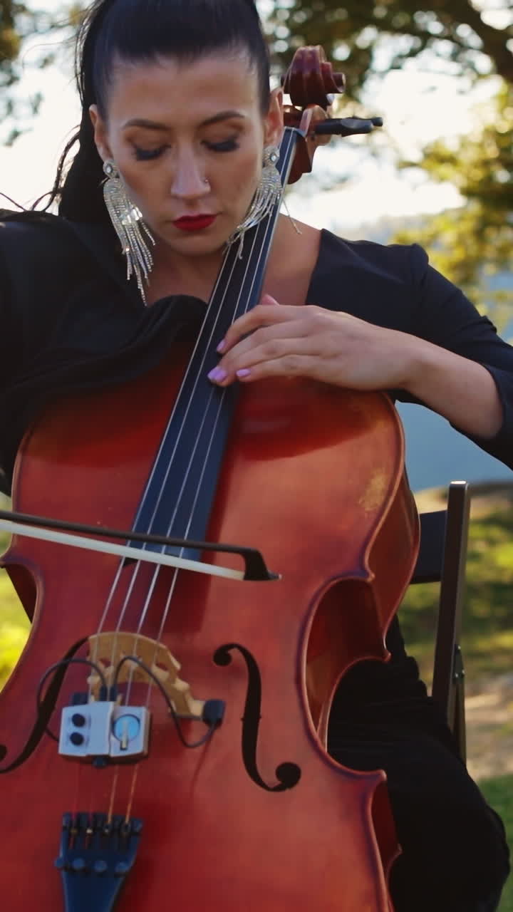 Dark-haired female musician focused on playing cello. Middle-aged woman in black dress plays instrument in the nature backdrop. Vertical video