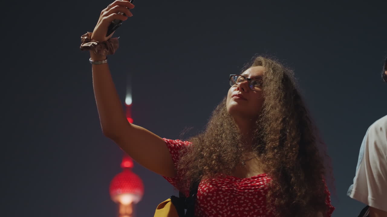 Donna con capelli ricci che si scatta un selfie notturno, abito rosso, smartphone tenuto in alto, torre illuminata che si erge dietro, le luci urbane brillano, posa spontanea con espressione gioiosa, atmosfera da influencer, celebrazione cittadina