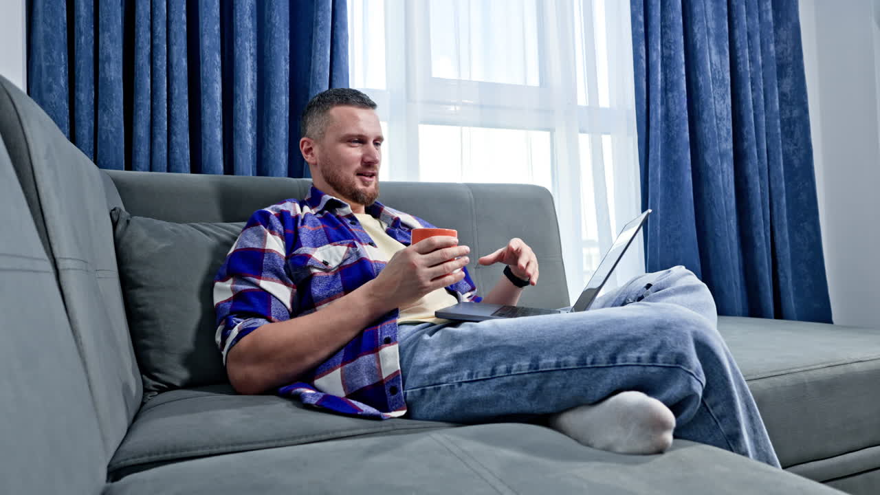 En mand sidder på en sofa med en kop i hånden, mens han taler via en bærbar computer i en lys stue med blå gardiner om dagen